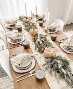 If simplicity is your style, this minimalist tablescape setup idea is for you. Soft cream and white tones dominate the table, with candles providing warmth. Add a garland of greenery and a few pastel-colored pumpkins to tie everything together in an understated yet elegant way.