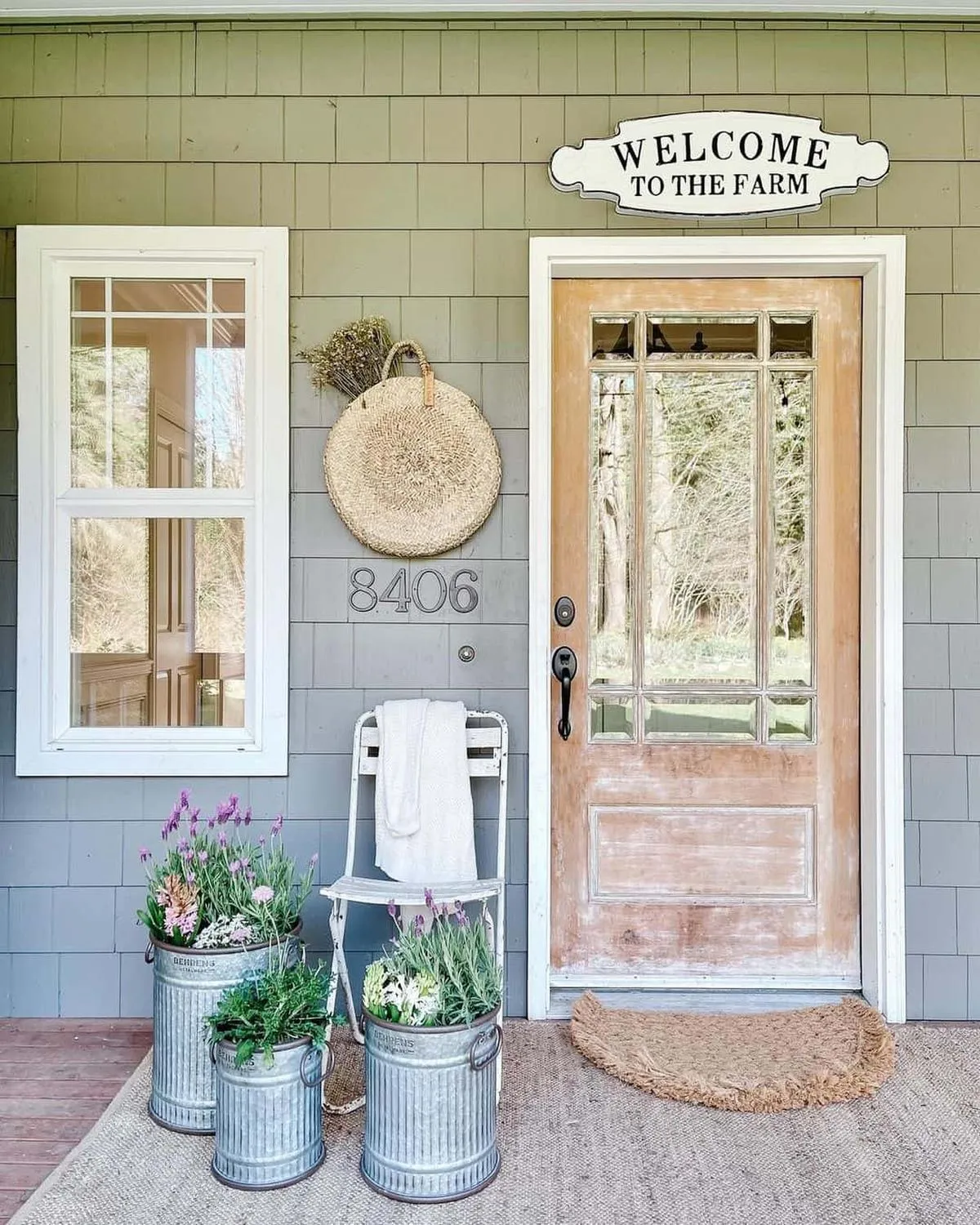 For lovers of understated elegance, this farmhouse-style porch features galvanized planters brimming with lavender and soft greenery. The vintage-inspired door and welcoming mat invite guests into a serene and charming space. Adding a woven basket and a simple white chair enhances the overall rustic aesthetic, making this decor effortlessly timeless. 