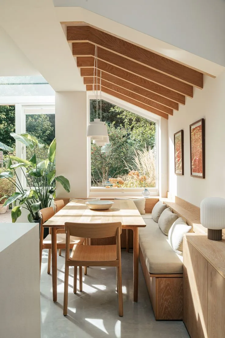 Organic-inspired corner dining nook featuring a sloped wooden ceiling, soft tones, and indoor plants for a nature-infused ambiance.