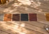 Deck Stain Colors: I Tested 7 Shades So You Don’t Have To Seven deck stain color samples in a row on a wooden pine board, ranging from gray to dark brown, with a paintbrush and stain can nearby.