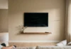 Minimalist TV Wall Design: 5 Approaches That Work in Real Living Rooms Extremely minimal TV wall with a flat-screen mounted on warm white plaster, no cables and no decor — just the screen, afternoon light, and a small plant on the floor.